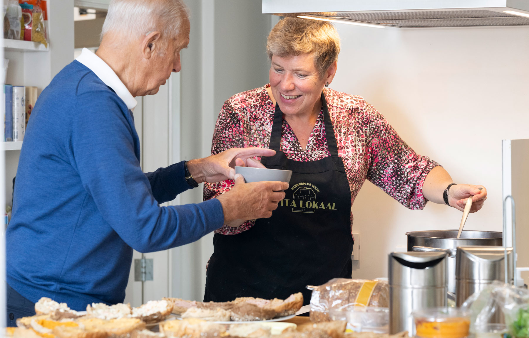 Een afbeelding van een vrouw die een kopje soep geeft aan een oudere man bij Evita Lokaal